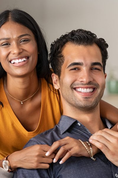 Smiling indian woman hugging her husband on the couch from behind at home. Loving middle eastern couple looking at camera with big grin in the living room. Portrait of laughing girl embracing handsome latin man on sofa.